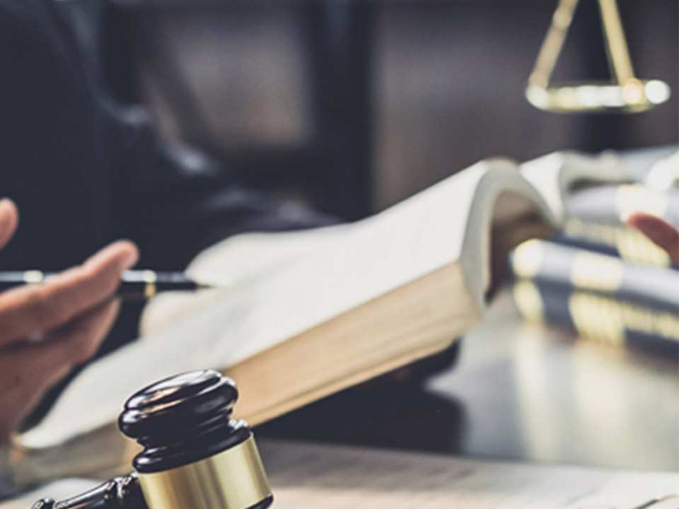 AsesoresJDX-Portafolio-Imagen Primer plano de un mazo de juez sobre una mesa mientras dos personas discuten documentos legales en segundo plano. Se ven libros y un portapapeles.