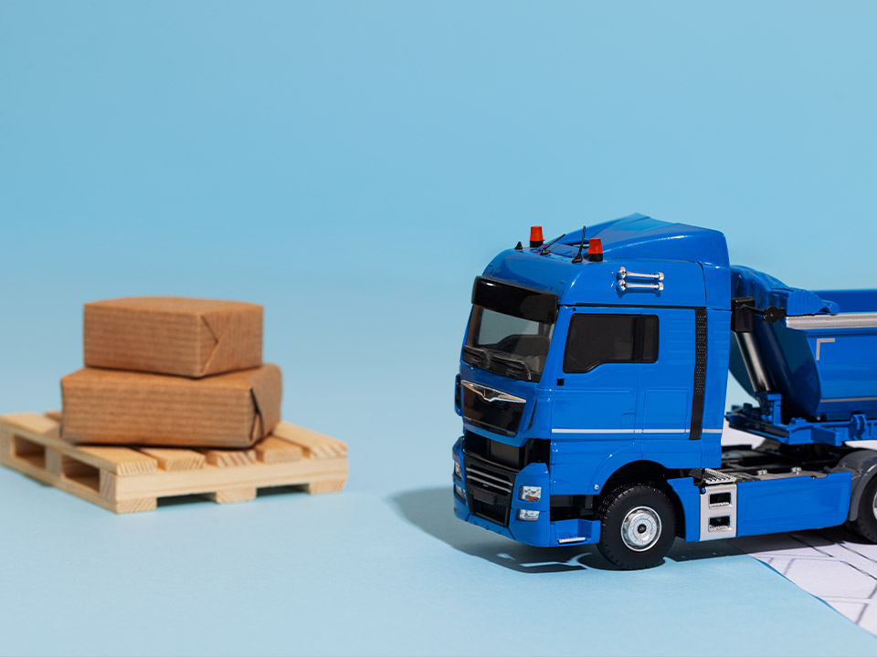 Anhanger-Portafolio-Truck-2 Camión azul de juguete junto a una pequeña tarima con paquetes marrones, en una representación de transporte y logística en miniatura.