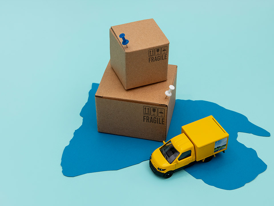 Anhanger-Portafolio-Truck-3 Escena minimalista con dos cajas de cartón marcadas como frágiles y una camioneta amarilla de juguete, colocadas sobre un mapa estilizado en tonos azules.