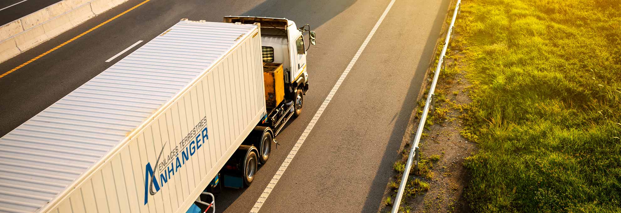 Anhanger-Portafolio-Truck-4 Camión de carga de la empresa Anhänger conduciendo por una autopista al atardecer, con el contenedor blanco y el logotipo visible en un entorno de carretera despejada.