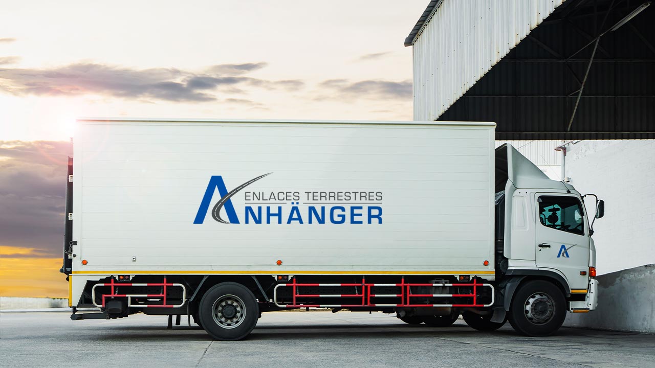 Anhanger-Portafolio-Truck Camión de la empresa Anhänger estacionado frente a un almacén o hangar, con el logotipo de la empresa claramente visible en el lateral del vehículo.