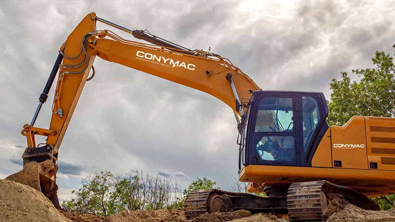 Conymac-Portafolio-Maquinaria-Marca Imagen de una excavadora naranja con el logotipo de CONYMAC en uso en un terreno de construcción, con un cielo nublado en el fondo.