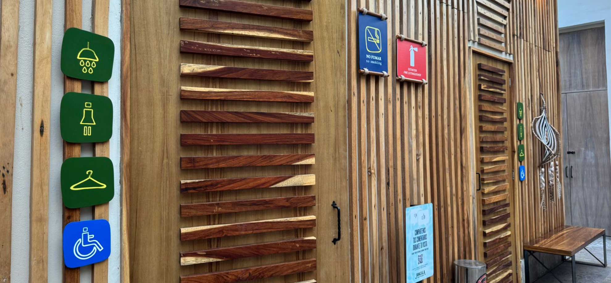 Jungala-Park-Portafolio-Bano Señalamientos sobre una pared de madera con íconos de ducha, vestidor y accesibilidad para discapacitados, junto a otras señales de 'No fumar' y extintor en Jungala Aqua Experience.