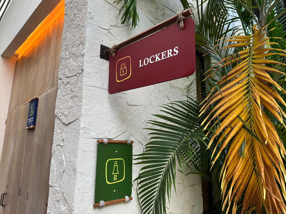Jungala-Park-Portafolio-Lockers-2 Señalética indicando la ubicación de los lockers, y señalamiento de baño de mujeres en Jungala Aqua Experience, montada sobre una estructura rústica, rodeada de vegetación tropical.