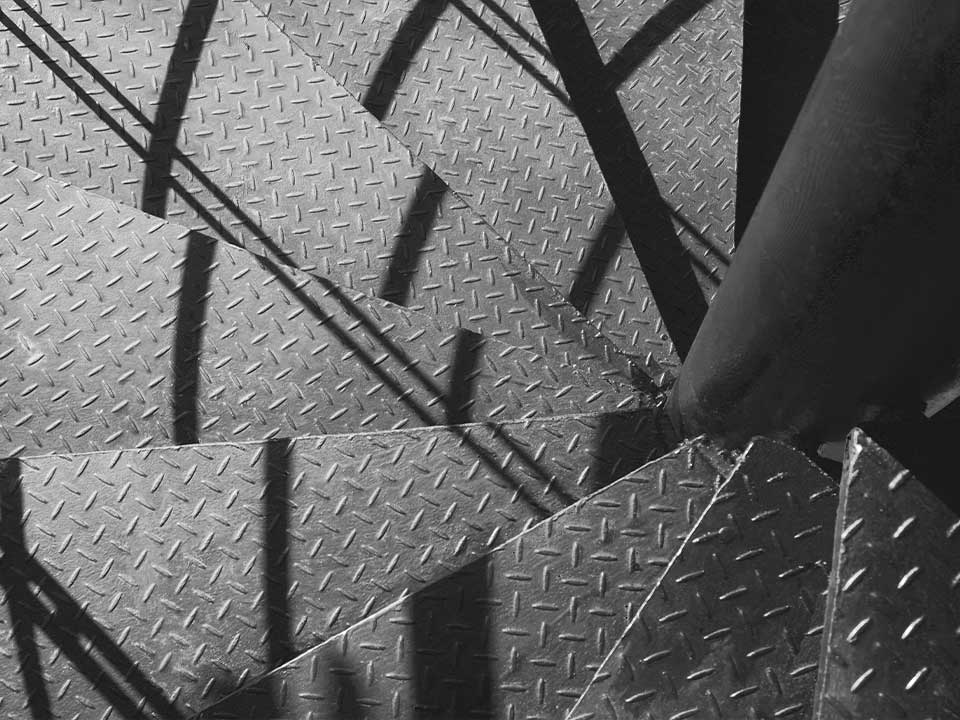 MetalyPunto-Portafolio-Metal-2 Imagen de una escalera metálica con un patrón de diamantes y sombras proyectadas que crean un contraste geométrico.