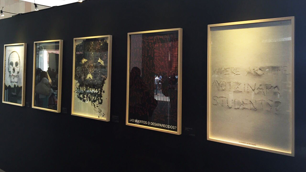 Exposición de cinco pósters gráficos enmarcados en una pared oscura, como parte de la bienal de carteles de Ayotzinapa. Los pósters reflejan preguntas y representaciones artísticas sobre la desaparición de los 43 estudiantes.