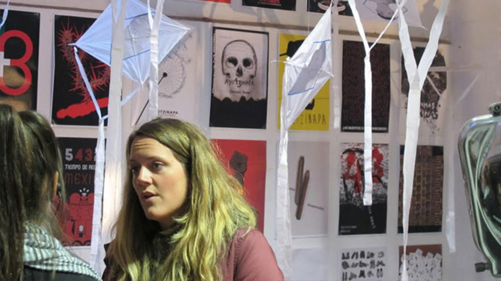Conversación entre dos personas dentro de la exposición bienal de carteles de Ayotzinapa. De fondo, cometas colgantes y carteles gráficos que presentan mensajes poderosos sobre temas sociales, como los estudiantes desaparecidos de Ayotzinapa.