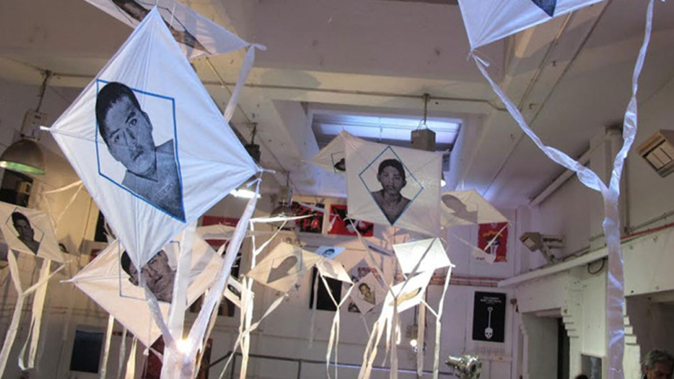 Exposición artística en la bienal de carteles de Ayotzinapa, con cometas colgando en el aire, cada una mostrando retratos de personas. La instalación está iluminada suavemente y las paredes están decoradas con pósters gráficos sobre temas sociales.
