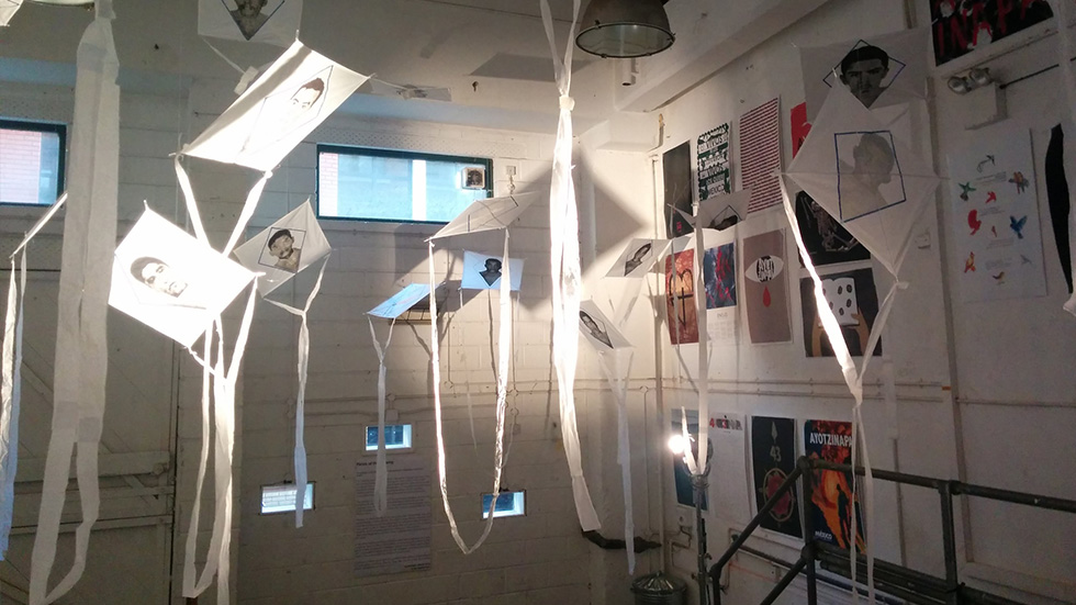 Instalación artística dentro de la galería, como parte de la exposición bienal de carteles de Ayotzinapa, con múltiples cometas de papel colgando del techo, cada una con retratos impresos. En las paredes, se pueden ver carteles de diferentes estilos gráficos.