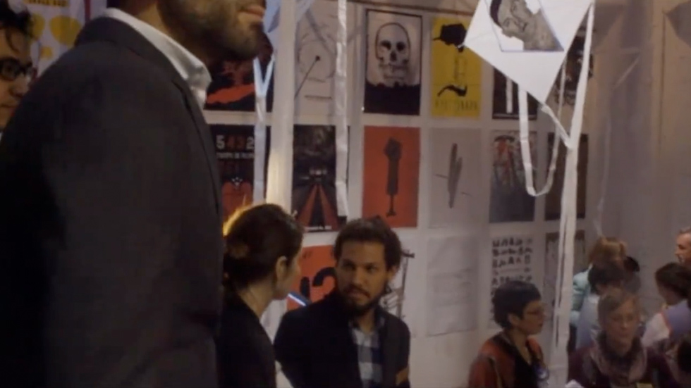 Personas dentro de la exposición bienal de carteles de Ayotzinapa, conversando y observando una instalación artística con cometas colgantes con retratos impresos. En el fondo, se aprecian carteles con temáticas de protesta social y arte conceptual.